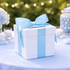 May include: A white gift box with a light blue ribbon and bow, set on a light blue tablecloth. The box has a slot on top. The background includes flowers, candles, and desserts, suggesting a celebration.