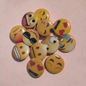 May include: A collection of yellow emoji face buttons with various expressions, including smiling, winking, and hearts for eyes. The buttons are round and have a glossy finish, with black and red accents. The buttons are scattered on a pink surface.