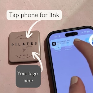 Op de afbeelding: Een vierkante beige NFC-tag met de tekst PILATES 1st STUDIO en een cirkelvormig grafisch ontwerp. Een smartphonescherm toont een Chrome NFC Tag-melding. Tekstoverlays bevatten Tap phone for link en Your logo here met bijbehorende pijlen.
