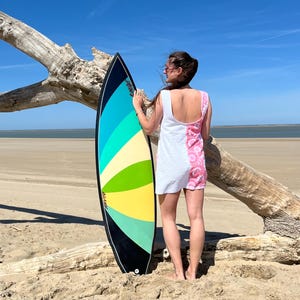 Peut inclure: Une planche de surf colorée avec un contour noir, présentant un design de rayures bleues, vertes et jaunes. La planche est tenue debout sur une plage de sable. Une robe blanche et rose à motifs est également visible.