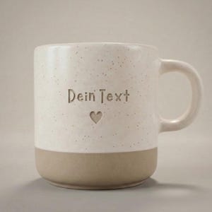 Könnte beinhalten: Cremefarbene Keramiktasse mit gesprenkelter Oberfläche und beigefarbenem Boden. Die Tasse trägt den geprägten Text "Dein Text" über einem kleinen Herz. Sie hat einen gebogenen Griff.