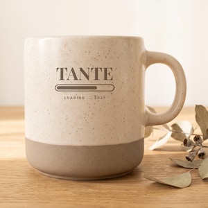 May include: A speckled cream-colored ceramic mug with a solid taupe base and a handle. The front features the dark brown text TANTE above a loading bar graphic and the text LOADING... 2027, printed in a clean, centered sans-serif and serif font.