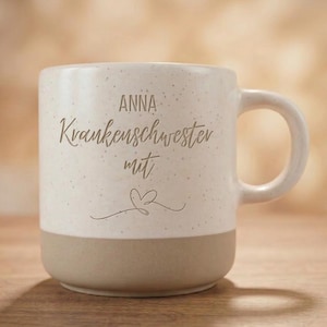 Könnte beinhalten: Ein gesprenkelter, cremefarbener Keramikbecher mit einem beigen Boden und einem Henkel auf der rechten Seite. Der Text "ANNA Krankenschwester mit" ist in Braun auf der Vorderseite aufgedruckt, ergänzt durch ein kleines, handgezeichnetes Herz.