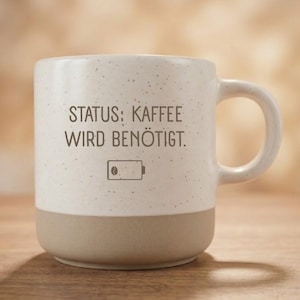 Könnte beinhalten: Ein Keramikbecher mit einem gesprenkelten, cremeweißen oberen Teil und einem hellbraunen Boden. Der Text "STATUS: KAFFEE WIRD BENÖTIGT." ist in Dunkelgrau aufgedruckt, ergänzt durch ein Symbol für einen fast leeren Akku mit einer Kaffeebohne.