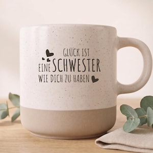 Könnte beinhalten: Ein gesprenkelter, cremeweißer Keramikbecher mit einem einfarbigen, hellbeigen Boden. Auf der Vorderseite befindet sich der schwarze Schriftzug GLÜCK IST EINE SCHWESTER WIE DICH ZU HABEN, ergänzt durch kleine schwarze Herzsymbole um den Text.