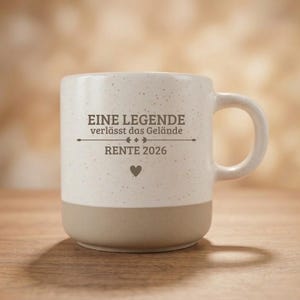 Könnte beinhalten: Ein gesprenkelter, cremeweißer Keramikbecher mit einem einfarbigen hellbraunen Boden. Auf der Vorderseite befindet sich der dunkelbraune Schriftzug EINE LEGENDE verlässt das Gelände, RENTE 2026 sowie ein kleines Herzsymbol unter dem Text. Mit Henkel.