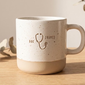 May include: A speckled off-white ceramic mug with a smooth tan base. The front features an engraved stethoscope icon centered between the text DOC on the left and [NAME] on the right. The mug has a rounded handle and a cylindrical body shape.