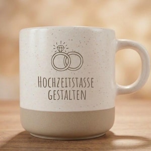 Könnte beinhalten: Eine cremefarbene und beige Keramiktasse mit Henkel. Auf der Tasse ist eine Zeichnung von zwei ineinandergreifenden Ringen, einer mit einem Diamanten, und der Schriftzug "HOCHZEITSTASSE GESTALTEN" abgebildet.