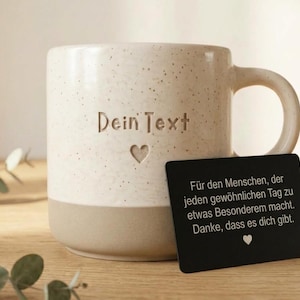 May include: A speckled cream and tan ceramic mug featuring the engraved text "Dein Text" above a small heart icon. A black rectangular card is placed in front of the mug with German text printed in white, ending with a small white heart icon.