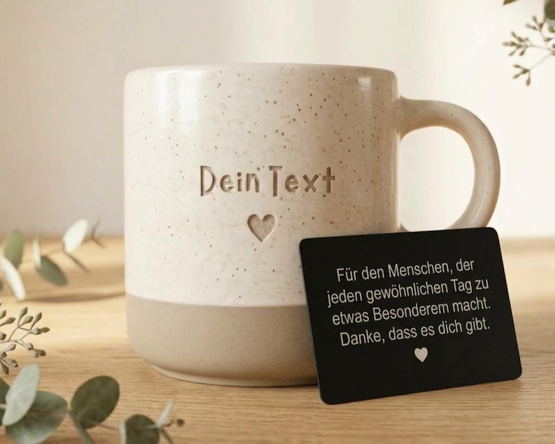 May include: A speckled cream and tan ceramic mug featuring the engraved text "Dein Text" above a small heart icon. A black rectangular card is placed in front of the mug with German text printed in white, ending with a small white heart icon.