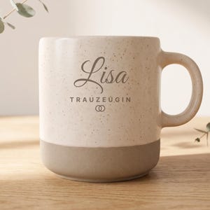 Könnte beinhalten: Eine Keramiktasse mit einem gesprenkelten cremefarbenen oberen Bereich und einem einfarbigen, matten grauen Boden. Auf der Vorderseite steht der Name Lisa in einer dunkelgrauen Schreibschrift, gefolgt vom Text TRAUZEUGIN und zwei Ringen.