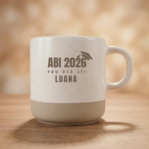 Könnte beinhalten: Ein gesprenkelter, cremeweißer Keramikbecher mit einem glatten, beigefarbenen Boden. Auf der Vorderseite steht in dunkelbrauner Schrift ABI 2026, YOU DID IT! und der Name LUANA, ergänzt durch eine kleine Grafik einer Doktorhut-Kappe über 2026.