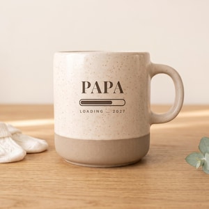 May include: A ceramic mug with a speckled cream upper section and a solid tan base. The front features dark brown text that reads PAPA, a loading bar graphic, and the text LOADING... 2027. The mug has a curved handle on the right side.