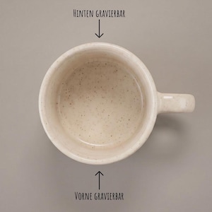 May include: A speckled off-white ceramic mug viewed from above with a handle on the right side. The text HINTEN GRAVIERBAR is above the rim with a downward arrow, and VORNE GRAVIERBAR is below the rim with an upward arrow pointing to the mug.