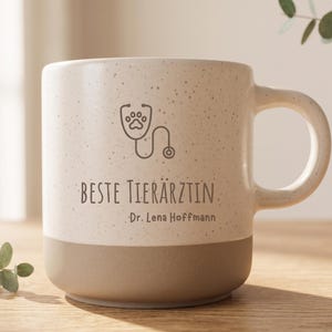 Op de afbeelding: Een gespikkelde, crèmekleurige en beige keramische mok met een donkerbruine afbeelding van een stethoscoop met een pootafdruk in het midden. De tekst "BESTE TIERÄRZTIN" staat boven de naam "Dr. Lena Hoffmann" in een donkerbruin lettertype.
