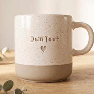May include: A ceramic mug with a speckled cream upper section and a solid matte beige base. The front features the engraved text "Dein Text" positioned above a small heart icon. The mug has a rounded handle and a smooth, cylindrical body.