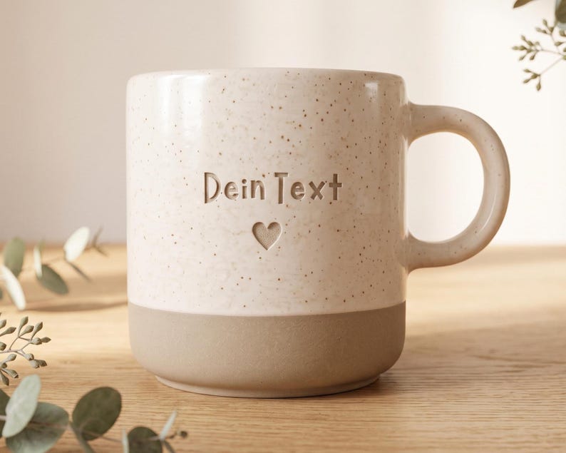 May include: A ceramic mug with a speckled cream upper section and a solid matte beige base. The front features the engraved text "Dein Text" positioned above a small heart icon. The mug has a rounded handle and a smooth, cylindrical body.
