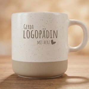 Può includere: Una tazza in ceramica con una parte superiore color crema maculata e una base marrone chiaro tinta unita. Il testo GERDA LOGOPÄDIN MIT HERZ con una piccola icona a forma di cuore è stampato in marrone sul davanti. La tazza ha un manico curvo.