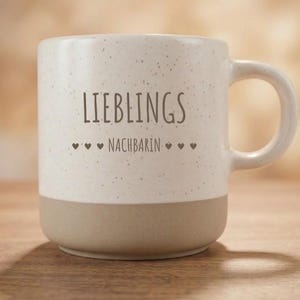 Könnte beinhalten: Ein Keramikbecher mit einem gesprenkelten cremefarbenen oberen Teil und einem einfarbigen hellbraunen Boden. Der Text LIEBLINGS NACHBARIN ist in Dunkelbraun auf der Vorderseite aufgedruckt, flankiert von kleinen Herzsymbolen neben dem Wort NACHBARIN.