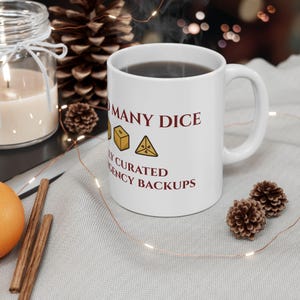May include: A white ceramic mug featuring the dark red text TOO MANY DICE and CAREFULLY CURATED EMERGENCY BACKUPS, with illustrations of a yellow six-sided die and a yellow four-sided die positioned between the two lines of text.