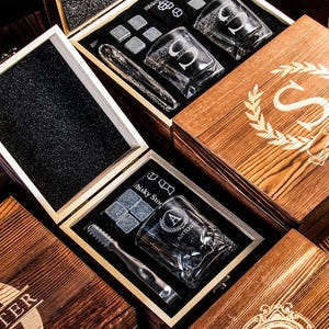 May include: Wooden gift boxes with engraved initials and designs. Each box contains a rocks glass, ice cubes, and tongs. The glasses are etched with letters. The boxes are made of wood with a natural finish.