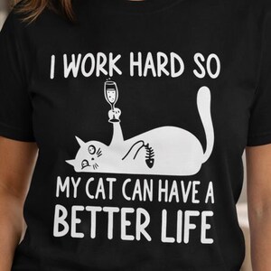 Puede incluir: Camiseta negra con el texto "I WORK HARD SO MY CAT CAN HAVE A BETTER LIFE". El gráfico presenta un gato blanco sosteniendo una copa de vino. La camiseta es de cuello redondo.