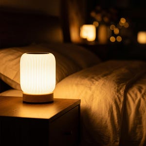 May include: A glowing, cylindrical bedside lamp with a ribbed, off-white shade sits on a wooden base. The lamp is on a dark wooden nightstand next to a bed with white bedding. The warm light creates a cozy atmosphere.