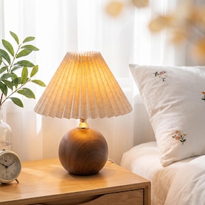 May include: A table lamp featuring a spherical dark wood base and a pleated, off-white conical fabric lampshade. The lamp emits a warm, soft glow, with a metallic gold-toned connector piece situated between the wooden base and the shade.