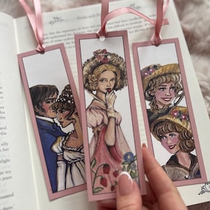Op de afbeelding: Drie rechthoekige bladwijzers met illustraties van vrouwen in historische kleding, elk met een roze rand en een roze lint. De bladwijzers liggen op een open boek. De illustraties zijn gedetailleerd.