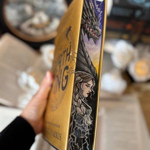 Op de afbeelding: Een goudkleurig boek met de titel "WORTH KING" en de naam van de auteur. De rand van het boek toont een draak en een vrouw met lang gevlochten haar. De draak is zwart en grijs met rode ogen. De vrouw heeft een verdrietige uitdrukking.