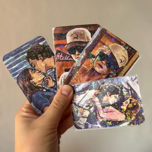 Op de afbeelding: Drie rechthoekige stickers met een holografische sterrenafwerking. Eén sticker toont een kussend stel, een andere een persoon met een pet en de tekst Stella, en de derde toont een omhelzend stel. Elke sticker heeft een levendig, geïllustreerd ontwerp.