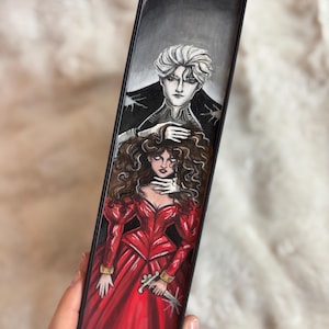 Peut inclure: Illustration peinte à la main sur un livre. L'œuvre représente un homme aux cheveux blancs et une femme en robe rouge. La femme tient un poignard. L'homme touche la tête de la femme. Le fond est noir et gris.
