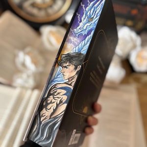 Puede incluir: Un libro de tapa dura con un borde pintado a medida que representa un dragón y una persona con tatuajes. El arte del borde utiliza tonos azules, blancos y morados. El lomo del libro es negro con letras doradas.