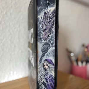 Puede incluir: Un libro de tapa dura con cubiertas negras y un canto pintado a mano que muestra un dragón morado sobre una figura de largo cabello blanco con una capa morada con capucha, sobre un fondo oscuro con rayos blancos y humo arremolinado.