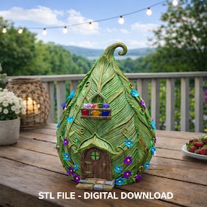 May include: A decorative fairy house in the shape of a leaf, with a green and gold finish. It has a small door, window boxes with colourful flowers, and is decorated with blue and purple flowers. The text "STL FILE - DIGITAL DOWNLOAD" is at the bottom.