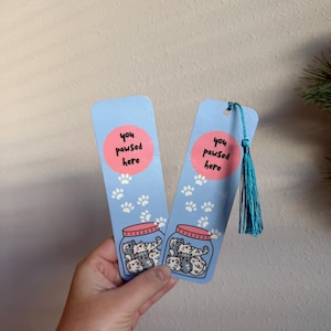 May include: Two blue bookmarks with a pink circle that says "you pawsed here". Each bookmark has a jar filled with cartoon cats and paw prints. One bookmark has a blue tassel.