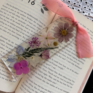 May include: A clear rectangular bookmark featuring embedded pressed flowers in shades of pink, purple, blue, and white. The bookmark is topped with a soft, light pink fabric ribbon tied in a knot, resting between the pages of an open book.