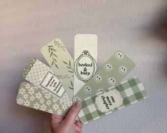 May include: A collection of six green and white bookmarks with various designs, including daisies, smiley faces, and gingham patterns. Each bookmark features text such as "booked & busy" and "one more chapter."