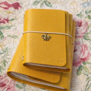May include: Mustard yellow leather journals with a bee charm and elastic closure. The journals are stacked on a floral patterned fabric. The initials "HY" are embossed on the front cover of the journal.