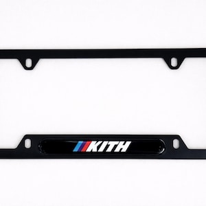 May include: Black license plate frame featuring the KITH logo in white, red, and blue. The frame is rectangular with rounded corners and mounting holes. The KITH logo is on a black bar.
