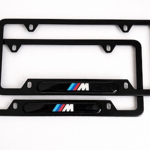 May include: Two black license plate frames with the BMW M logo in the center. The logo features the letters "M" in white, with blue, red, and light blue stripes. The frames are rectangular with rounded corners.