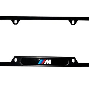Op de afbeelding: Zwart kentekenplaatraam met het BMW M-logo in het midden. Het logo toont de BMW-kleuren blauw, rood en wit. Het frame heeft een rechthoekige vorm met afgeronde hoeken en montagegaten.