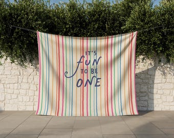 May include: A rectangular fabric backdrop featuring a vertical striped pattern in shades of pink, blue, yellow, and cream. The center of the fabric displays the dark blue text "IT'S FUN TO BE ONE" written in a combination of script and block lettering.