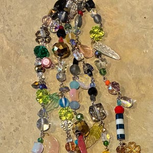 May include: A beaded keychain with a silver clasp. The keychain features strands of colorful beads, including clear, green, yellow, pink, and iridescent beads. Charms include a four-leaf clover, a lighthouse, and leaf-shaped beads.