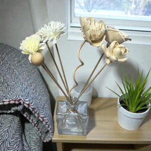 May include: A clear glass square diffuser bottle holds several decorative flower reeds. The reeds are topped with white and cream-colored artificial flowers. A small potted plant sits to the right on a light wood table.