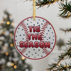 May include: A round Christmas ornament with a baseball theme. The ornament features red stitching, blue stars, and the text "TIS THE SEASON" in red. It hangs from a gold ribbon, perfect for holiday decor.