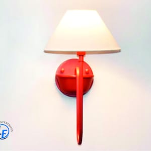 May include: A red wall sconce with a white lampshade. The sconce has a circular base and a long, curved arm. The lampshade is conical. The sconce is mounted on a white wall. The sconce is a decorative lighting fixture.