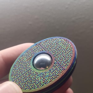 Puede incluir: Un fidget spinner multicolor con una superficie texturizada y un rodamiento de bolas plateado en el centro. El spinner tiene un degradado arcoíris alrededor de los bordes y un patrón tejido en la superficie.