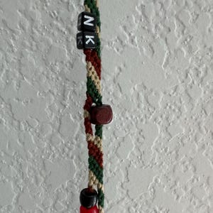 May include: A macrame keychain with a braided design in green, brown, and beige. The keychain features black letter beads spelling "NK", a wooden bead, and two smaller beads in black and red. The background is a textured white wall.