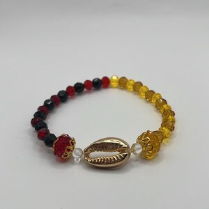 May include: A beaded bracelet featuring a central gold-toned cowrie shell charm flanked by clear beads and gold-toned filigree caps. The band consists of faceted black and red beads on one side and faceted yellow beads on the other side.